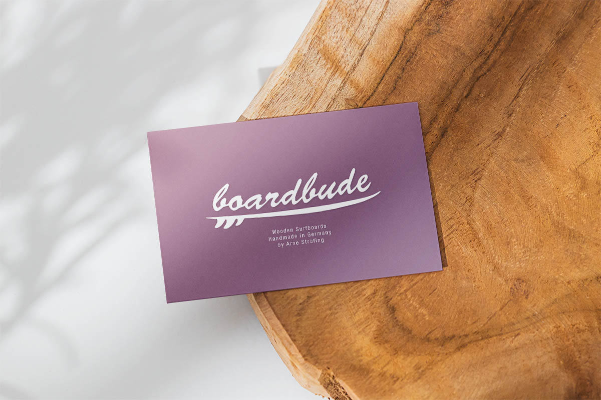  auf einem Holzbrett liegt die lilane Visitenkarte, das Logo Boardbude ist in weiß gehalten