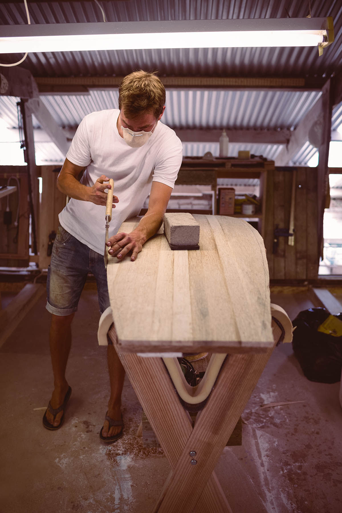 Der Shaper ist in seiner Werkstatt am arbeiten. Vor ihm liegt ein Board und in der Hand hält er eine Säge